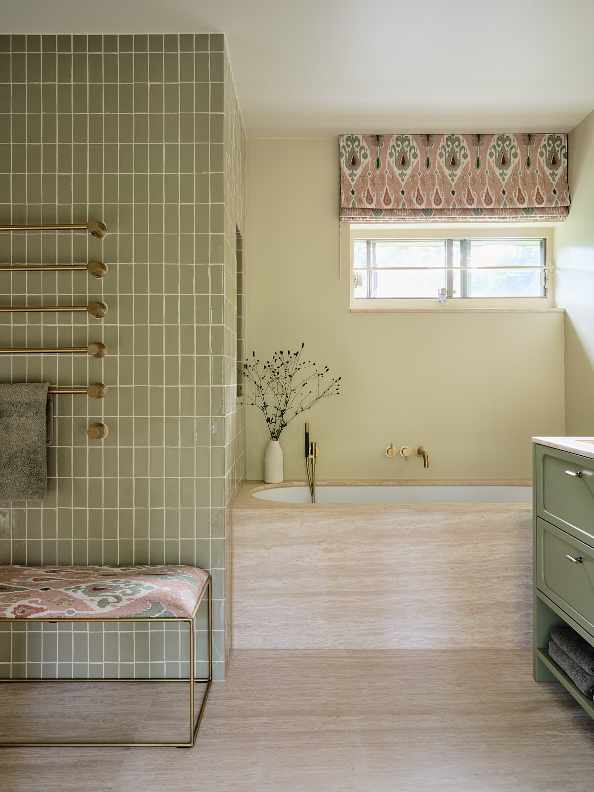 light green bathroom with travertine built-in bath, with green tiles, towel rail, bench seat and patterned roman blinds