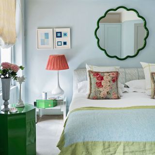 Master bedroom with cream carpet, pale blue walls and double bed with light blue blanket and vintage cushion with floral pattern, lamp with red lampshade on mirrored beside table