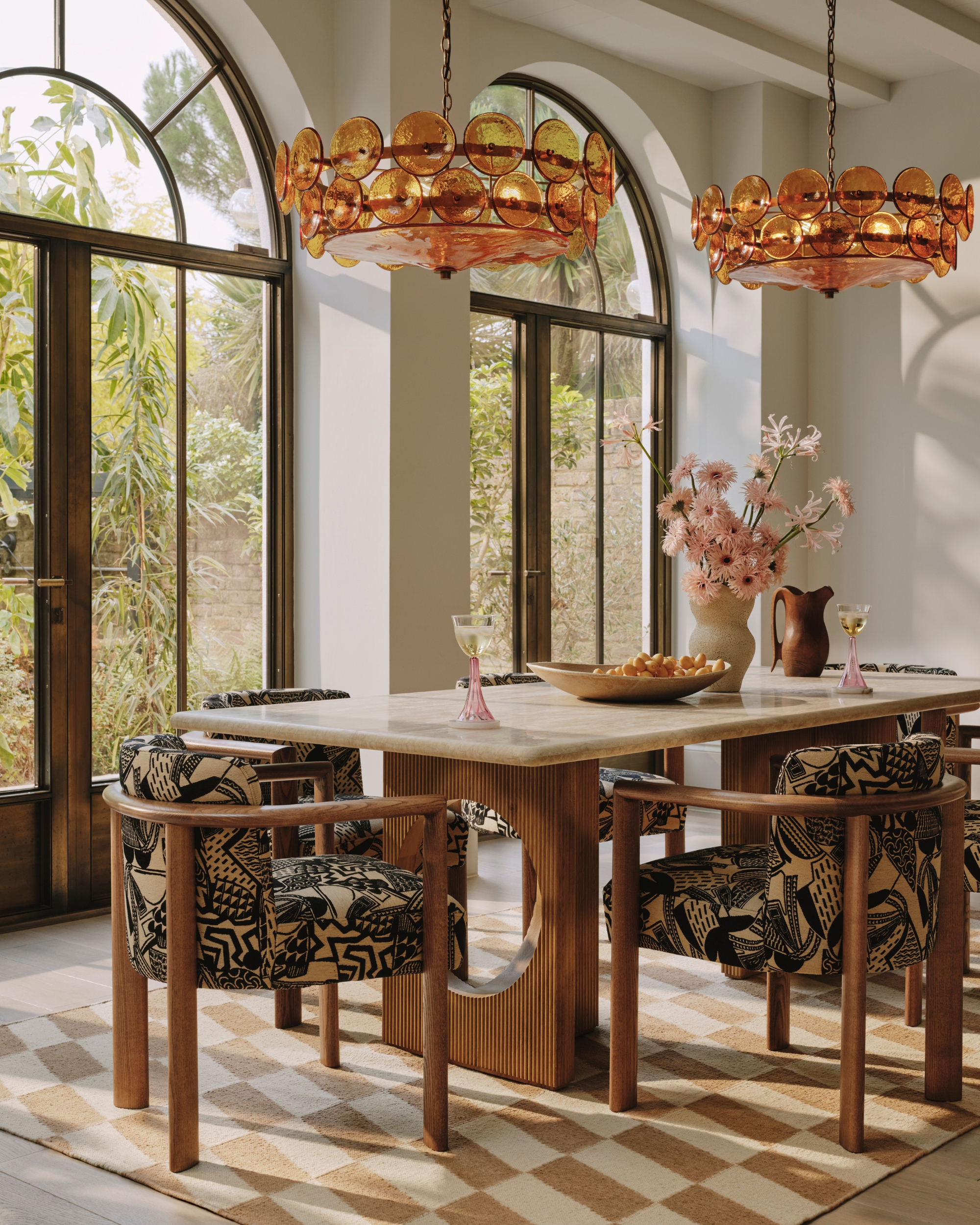 Image of a formal dining room with white walls, tall ceilings, and large floor-to-ceiling windows. The coffee table is wooden and marble with patterned seats all around it.