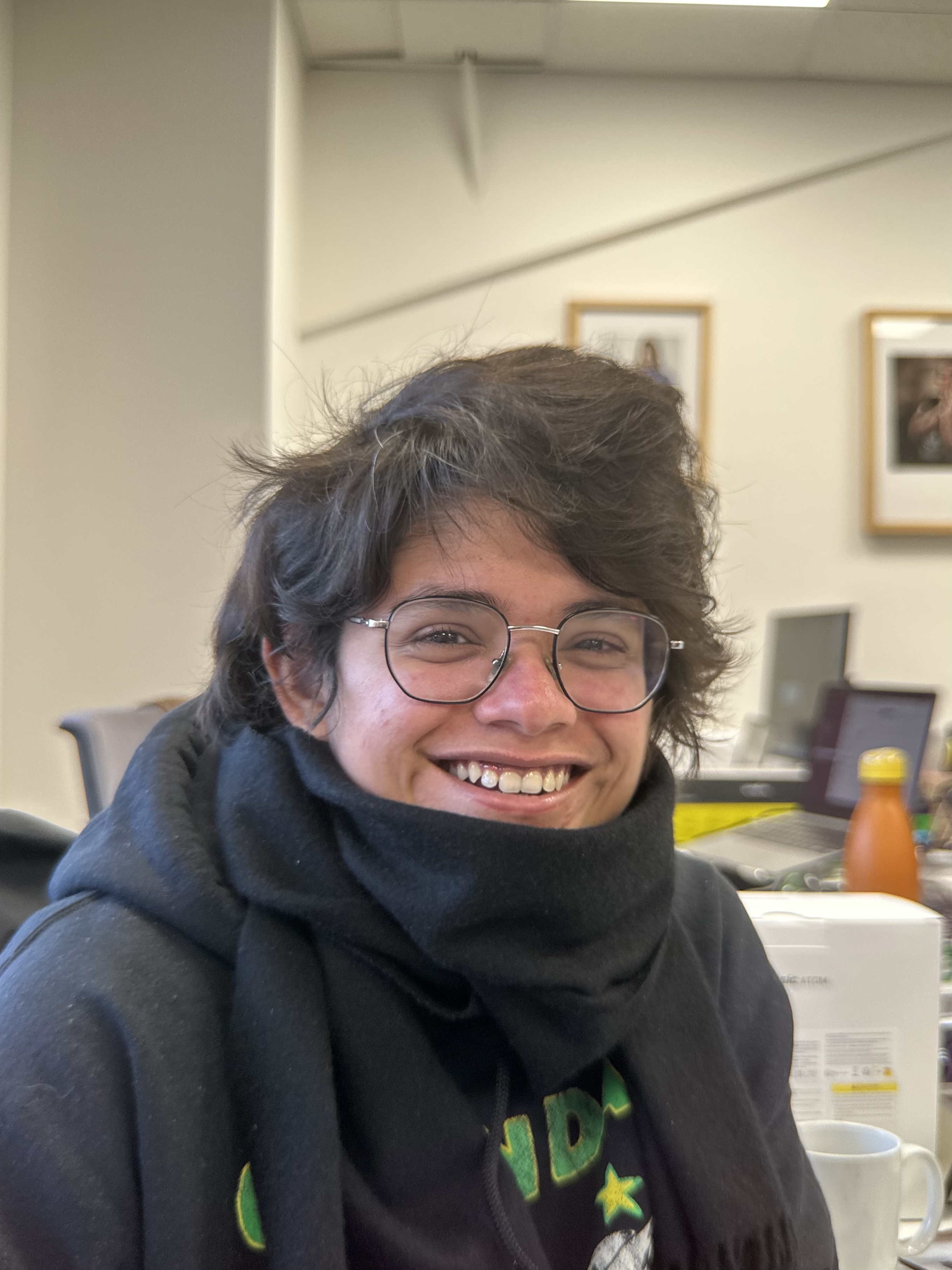 A photo of a person smiling in an office, with a black scarf around their neck and glasses.