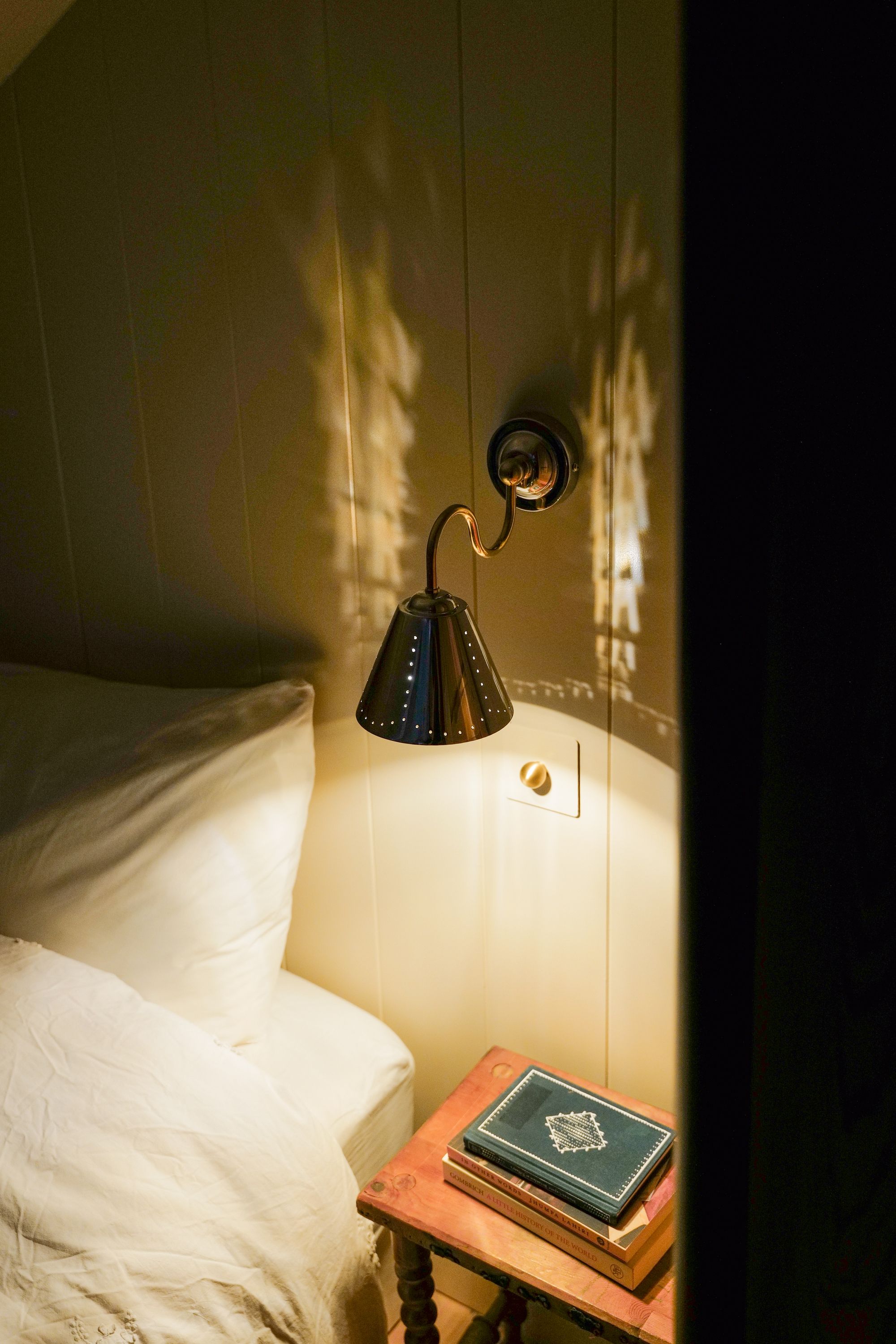 Image of a silver scone light with perforations in the shade. The light is turned on, and the room is dark. There is a minimalist but cozy white bed in view.