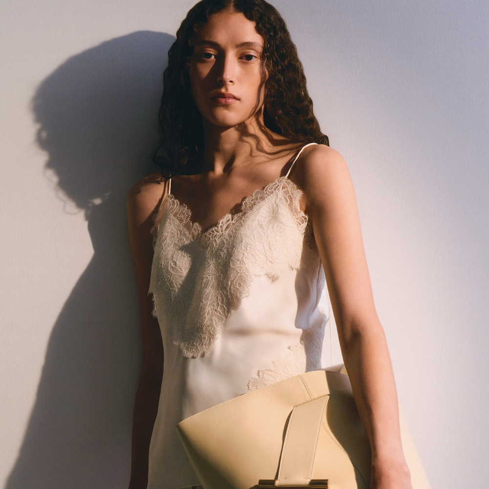 Model leaning agains a wall in soft lighting wearing a white lacey slip dress and holding a cream bag sold at Harvey Nichols