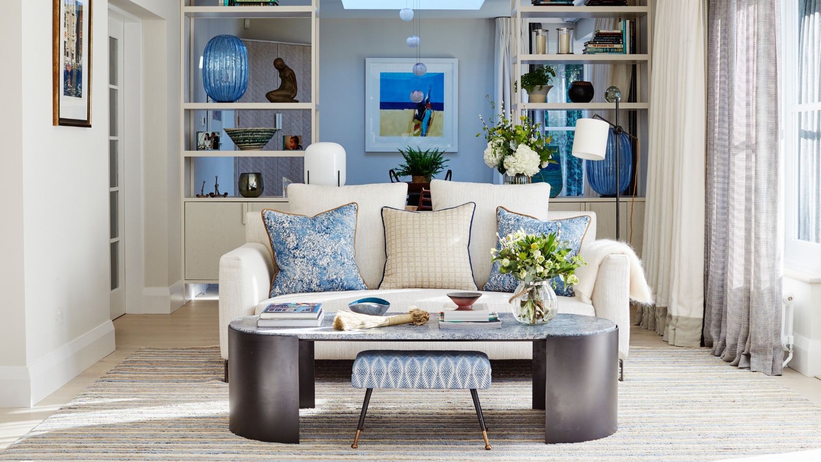A bright living room with an oval black and grey coffee table and small white sofa with blue and white cushions in the centre, with white walls with white display units, and pale grey rug. To the right is a large window with layered brown sheer curtains and white curtains.