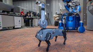 a four-legged dog-shaped robot with googly eyes and a thick antenna serving as a "head", inside a lab with equipment and computers