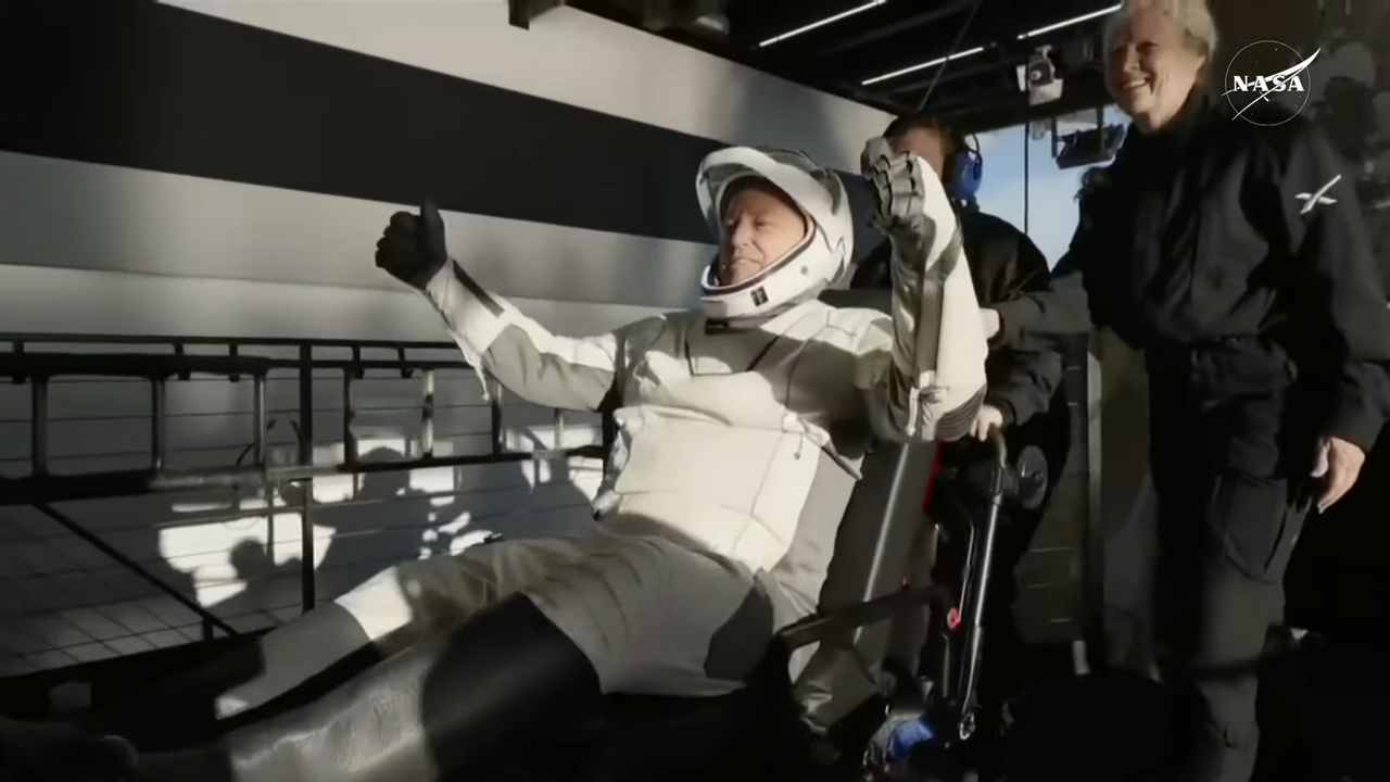 A male NASA astronaut in a white and black spacesuit raises both arms in triumph after leaving his Crew-9 spacecraft on stretcher