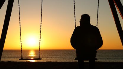 A man sits on a swing facing the sunset at the beach with an empty swing beside him.