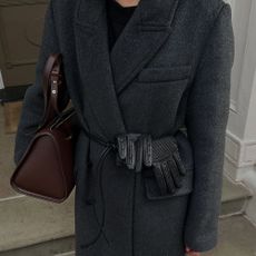 : @_santinaharrison wearing a dark grey boxy wool coat with a black crew-neck jumper, black leather cord belt, black leather gloves and brown leather bowling bag