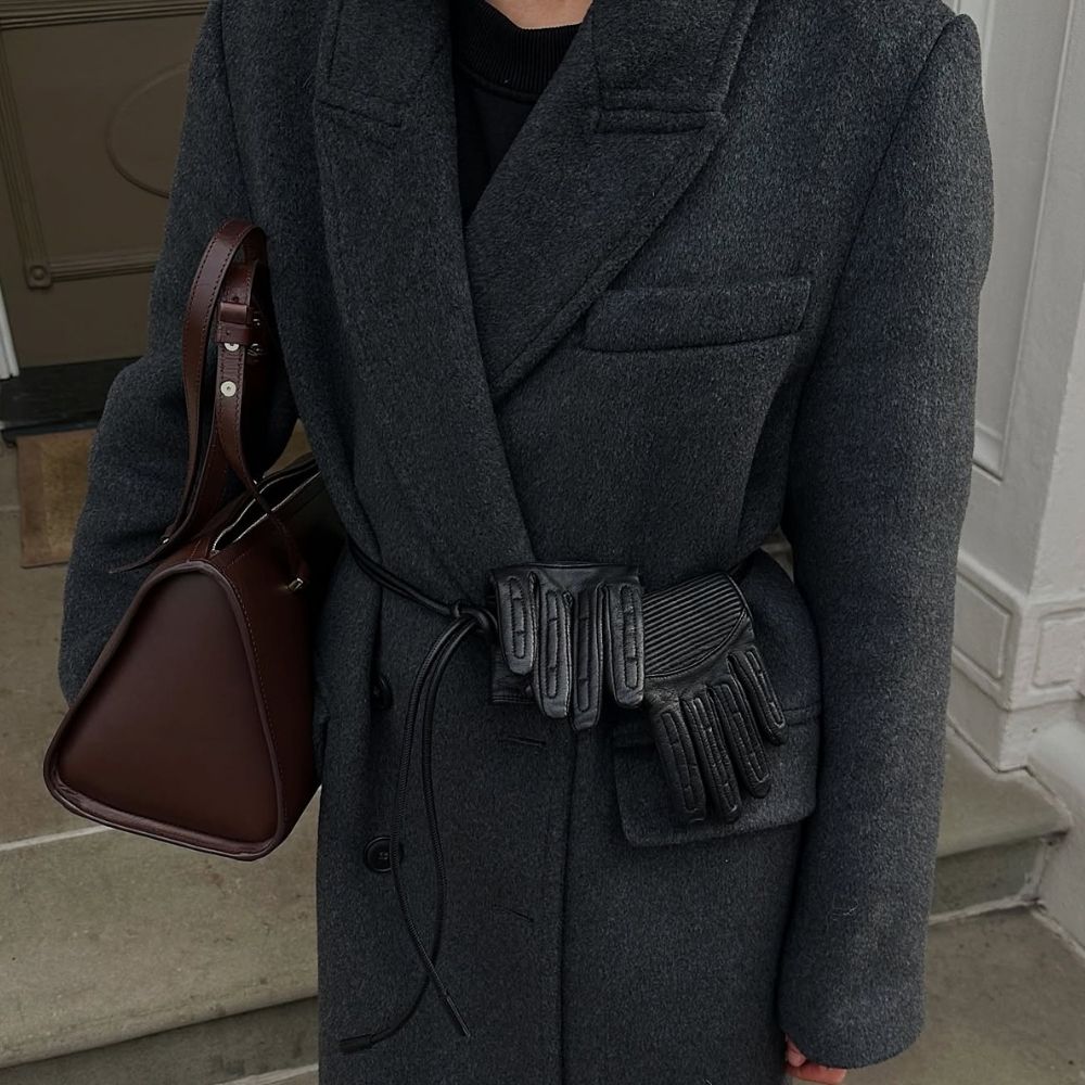 : @_santinaharrison wearing a dark grey boxy wool coat with a black crew-neck jumper, black leather cord belt, black leather gloves and brown leather bowling bag