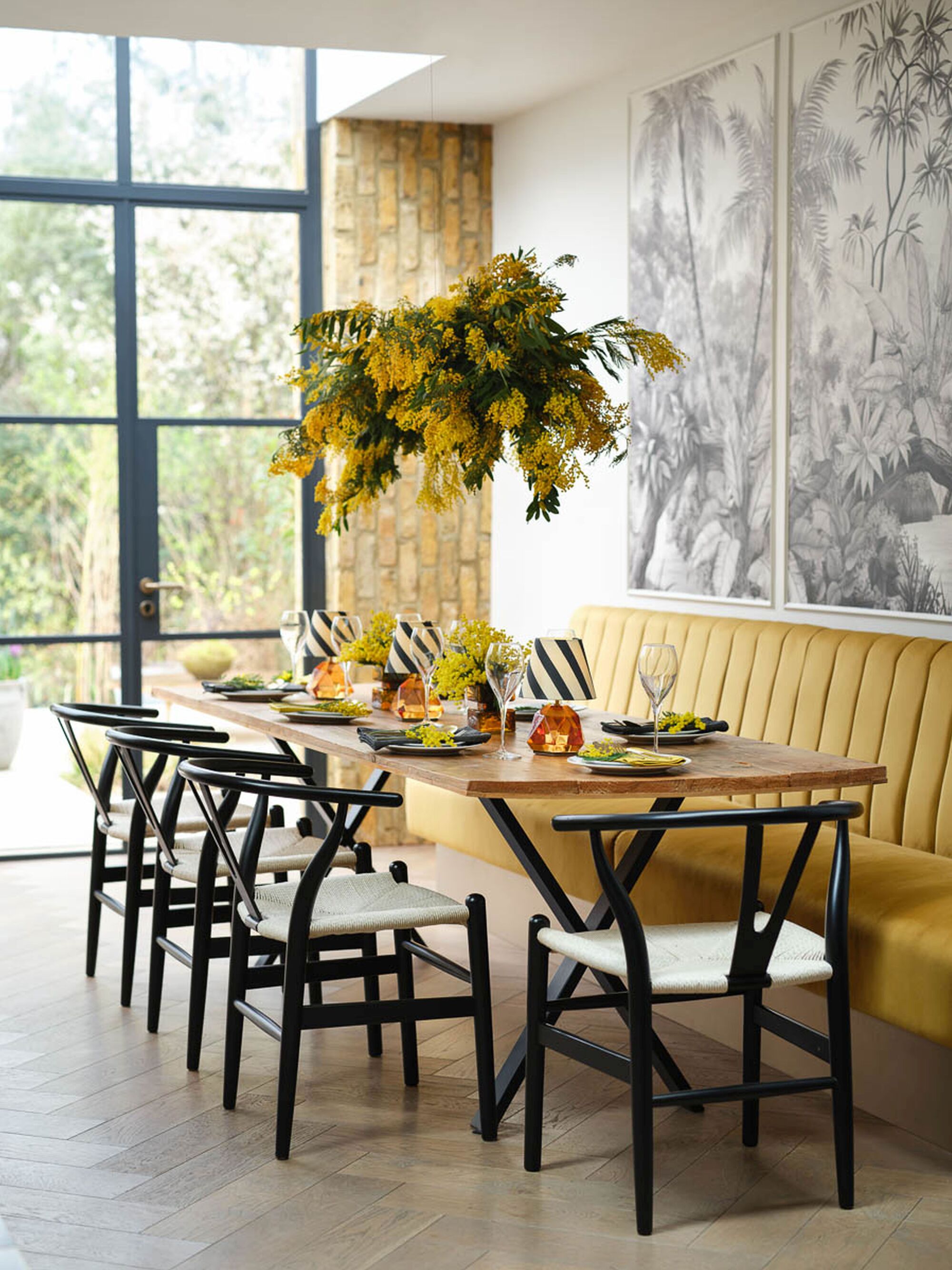 table with mustard banquette seat and mural wall, wishbone chairs, and florals hanging overhead, and table set with wine glasses, plates, and portable lamps