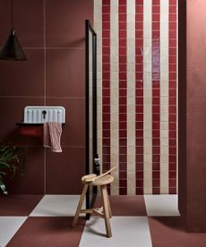 A cherry red bathroom with white and red tiles on the wall and on the floor, a black shower door, and a wooden stool with a brrush and a black bottle on top of it