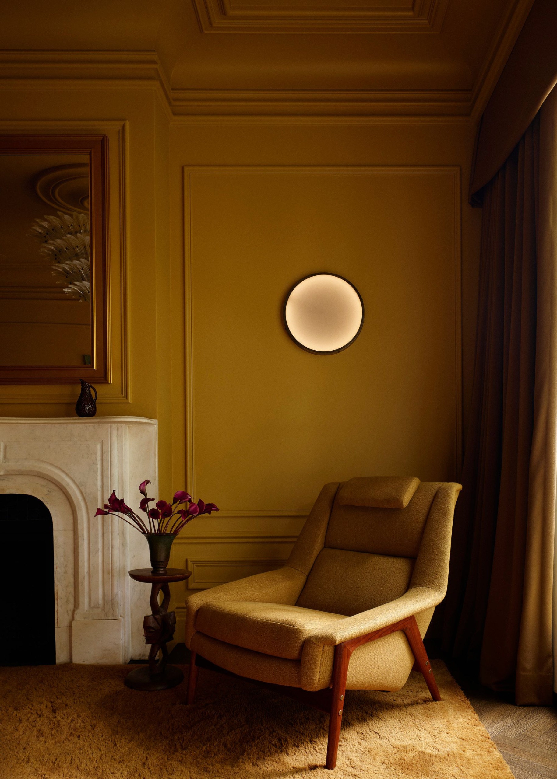 A modern bedroom with ochre walls and decorative moldings, a marble fireplace, a textured caramel-colored rug, and an ochre armchair.