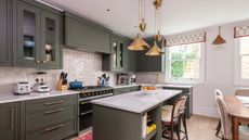 A modern kitchen with khaki green cabinetry, island and brass colored overhead kitchen island pendant lighting decor