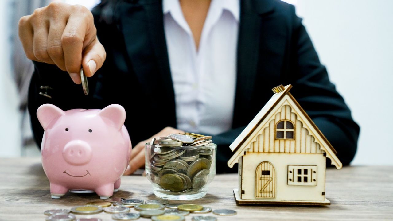 Person putting money into piggy bank next to jar of coins and model house