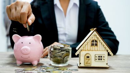 Person putting money into piggy bank next to jar of coins and model house