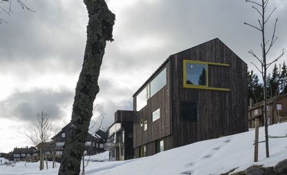 Exterior view of the Oslo family house by STA - the house features a dark wood exterior, windows with yellow frames and it is surrounded by other houses, trees and snow