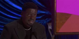 Daniel Kaluuya giving a speech on the Oscar stage dressed in a black jacket with a black shirt underneath.