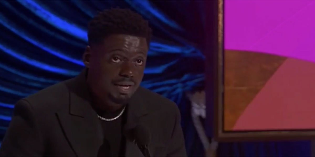 Daniel Kaluuya giving a speech on the Oscar stage dressed in a black jacket with a black shirt underneath.