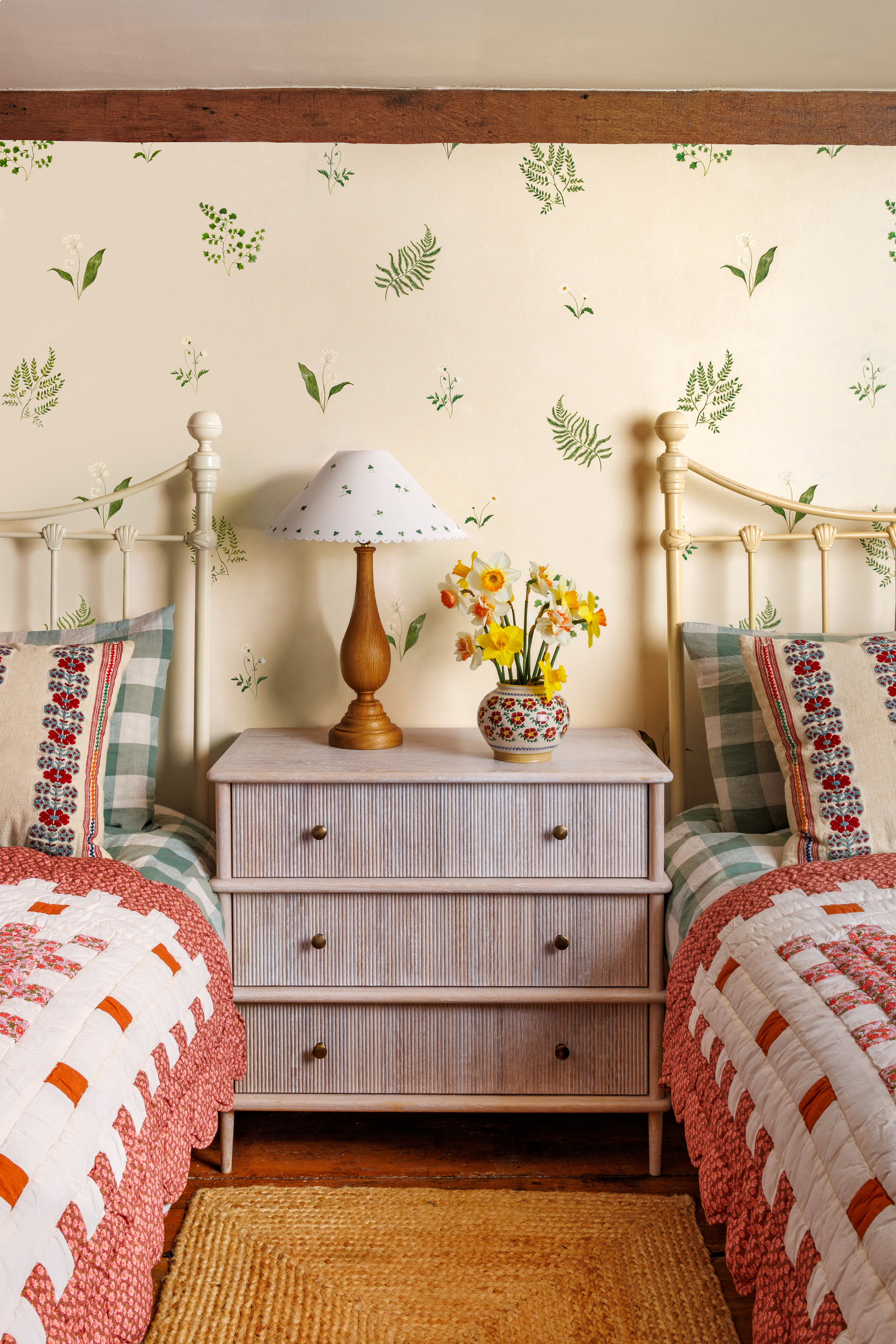 Image of a cottagecore-style bedroom with two twin beds that have pink and white quilts. There is a chest of drawers between the beds with a lamp that has a shade with drawings on it. The wallpaper is hand-painted.