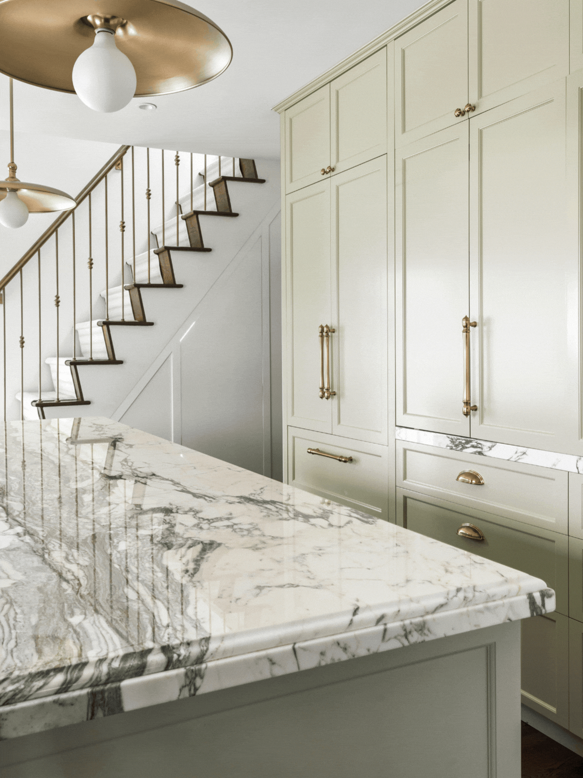 GIF of a cabinet opening in a light sage green kitchen to reveal a coffee nook that has a coffee machine, mugs, and bowls within it. There is a large marble island in the foreground.