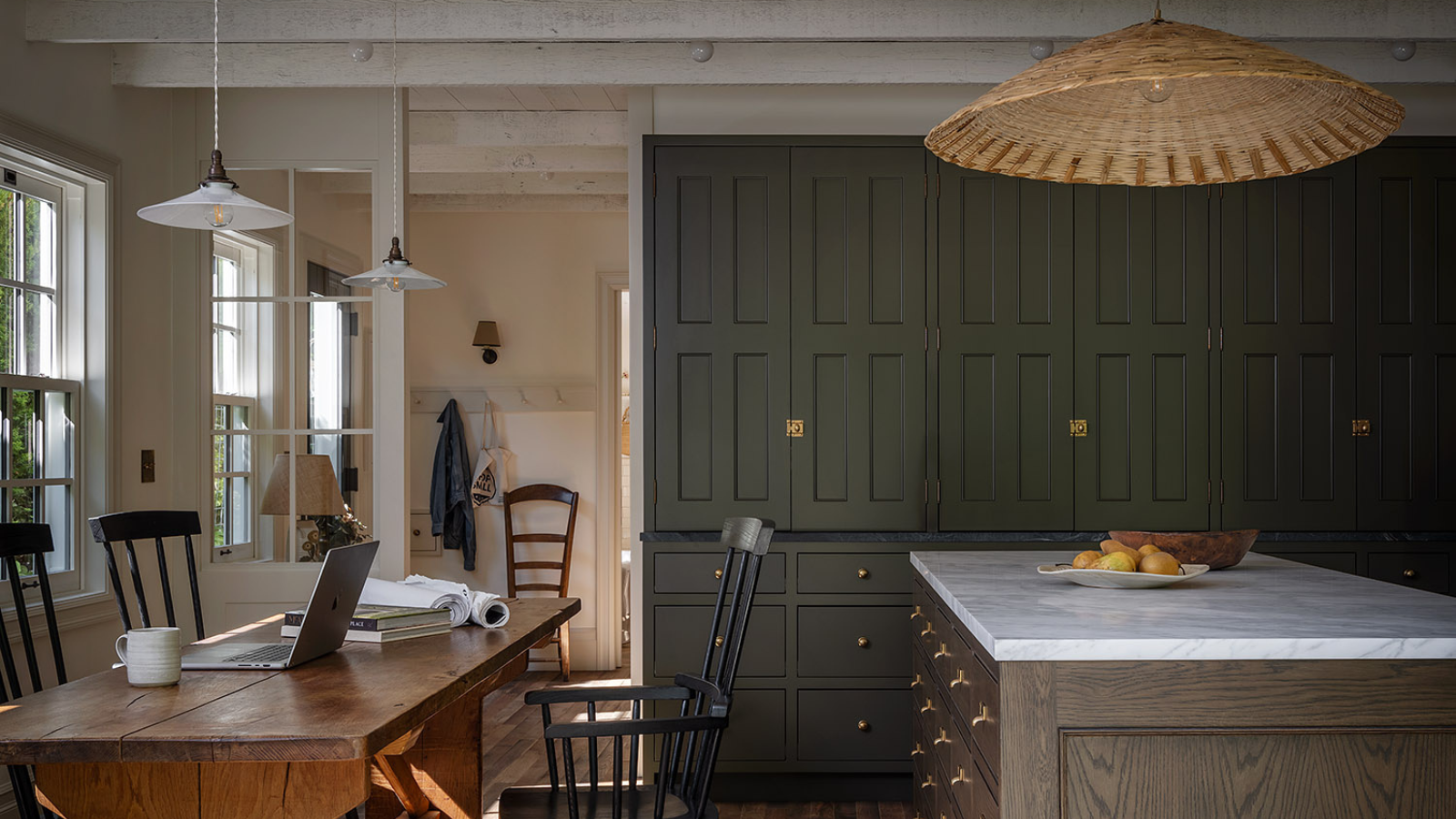 a rustic kitchen with a large wooden table and several chairs, a kitchen island with a marble countertop, a wall of cabinets painted in a deep green color, and a large rattan pendant lamp 