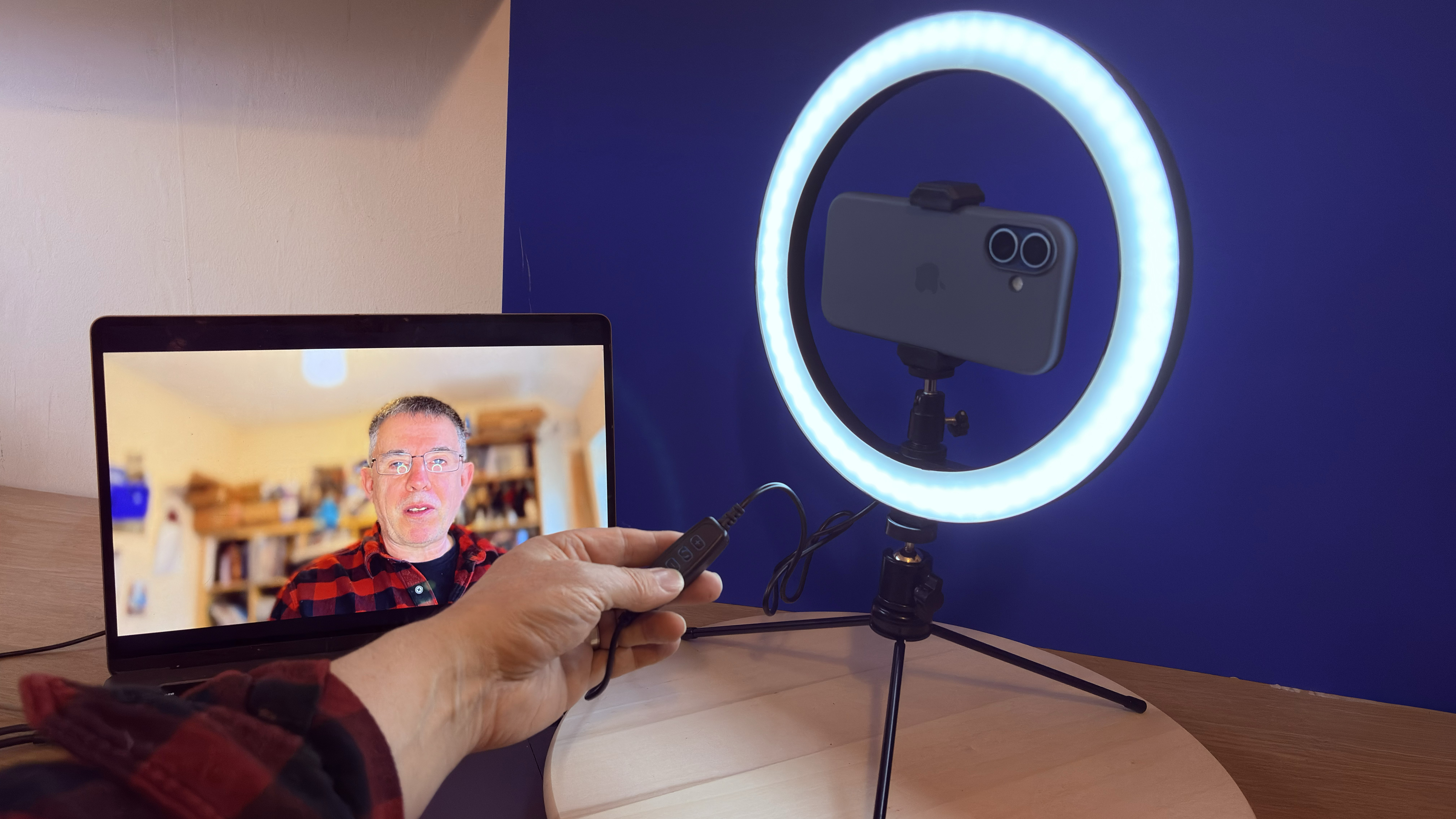 Shot of the AiXPi 10&amp;rdquo; Ring Light holding an iPhone 17. The feed from the iPhone&amp;rsquo;s camera is being displayed on a MacBook. A hand holds the LED&amp;rsquo;s control unit.