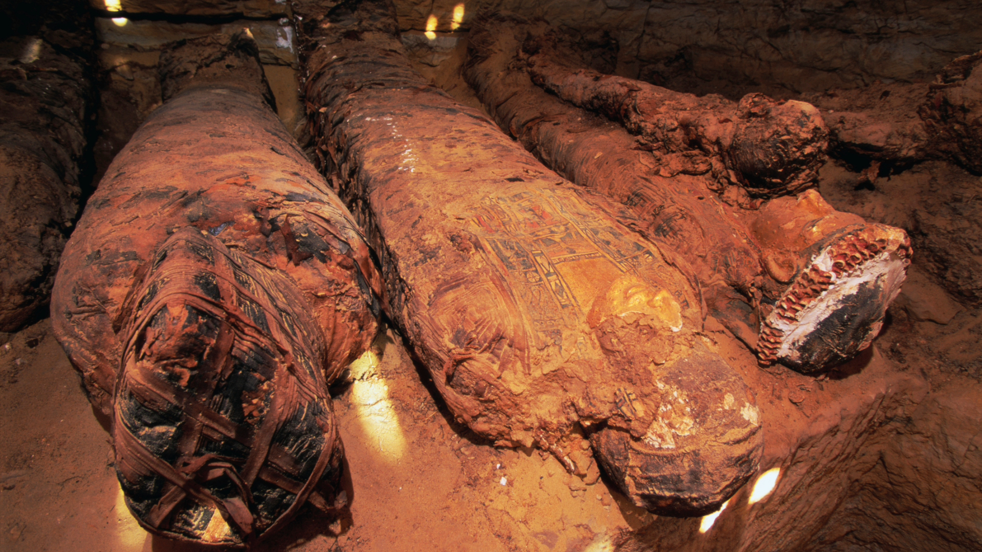 Three preserved mummies lie flat on the sands in an Egyptian tomb