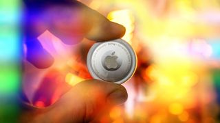 An Apple AirTag held between a thumb and forefinger against a blurry backdrop of bright colors.