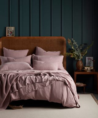 Dusky rose relaxed bedlinen and plentiful pillows, with cane headboard, and dark wood paneling on walls.