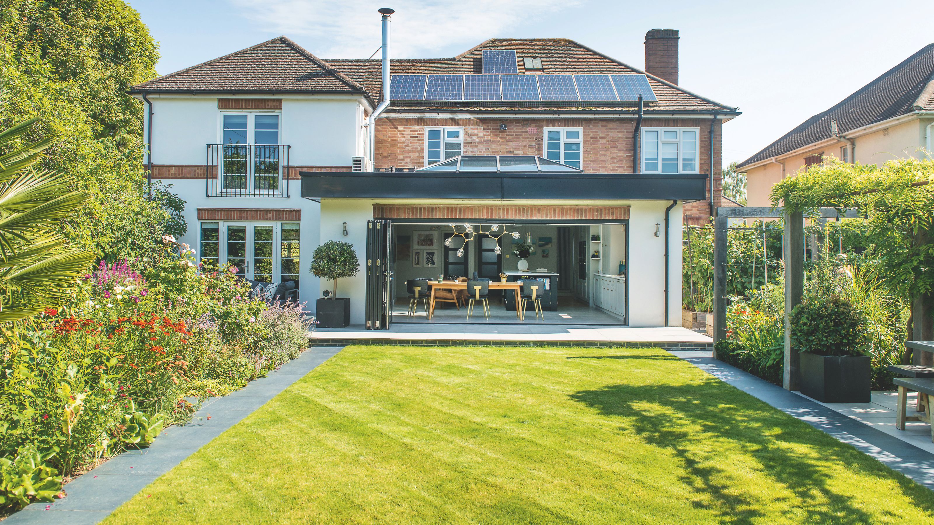 Back of a large house with an open dining room area opening up to a large lawn. There's a covered seating area on the right and established plants on the left