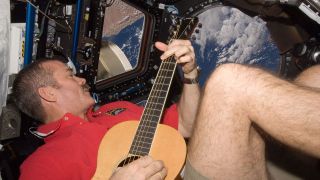 a man in a red shirt and shorts plays guitar while floating weightless in a cramped room full of windows. earth can be seen through the windows