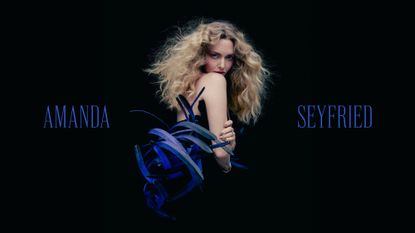 Amanda Seyfried stands in front of a black backdrop wearing a strapless black dress with blue feather detailing, and her blonde hair is styled curly. She is placed between her first and last name written in the same shade of blue.
