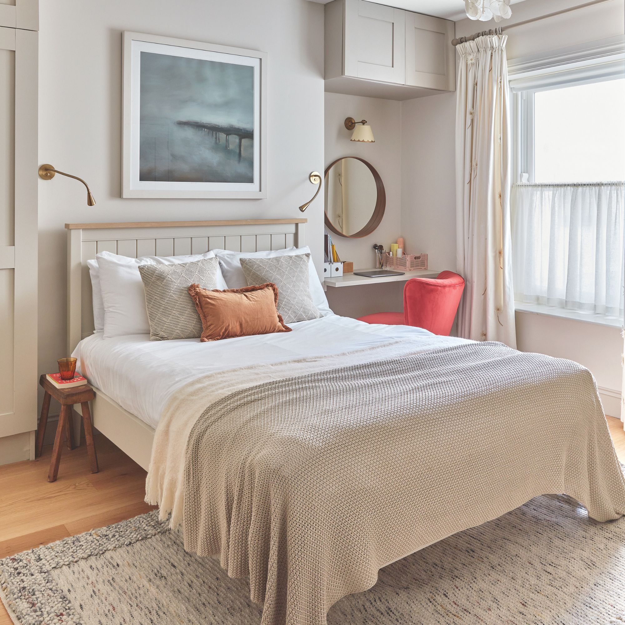 Neutral painted bedroom with layered bedding, a cream rug on the floor, and a bright red dressing table chair in front of dressing table