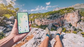 Person sitting on clifftop overlooking mountains holding smartphone with map locations open