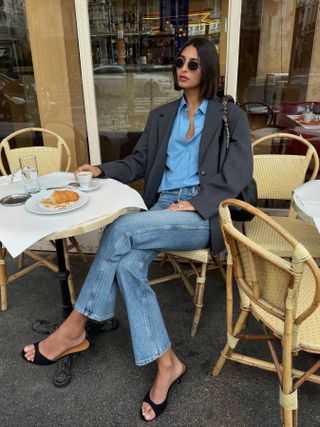 Fashion influencer and model Salomé Mory with a short bob haircut sitting outside a cafe in Paris wearing jeans, a blue button-down shirt, faded straight-leg jeans, and black open-toe heels with a Loewe bag over her shoulder.