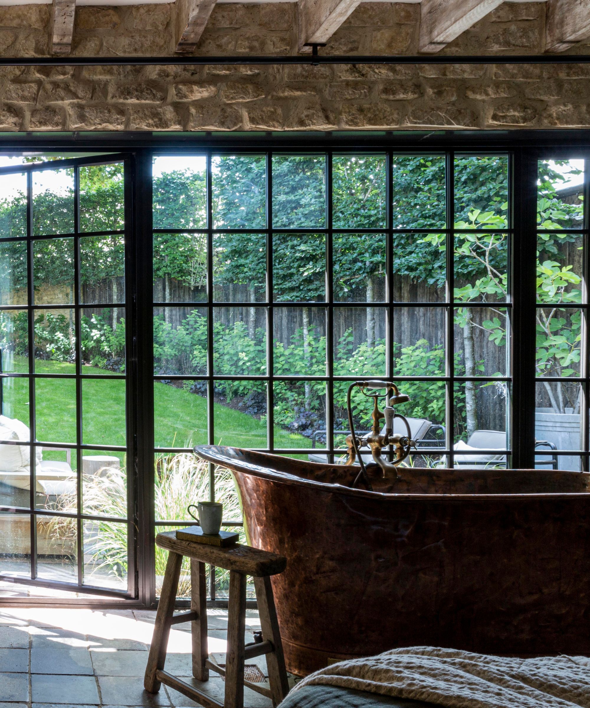 A bedroom with a copper bathtub in front of the window, styles with a wooden stool topped with a mug and book