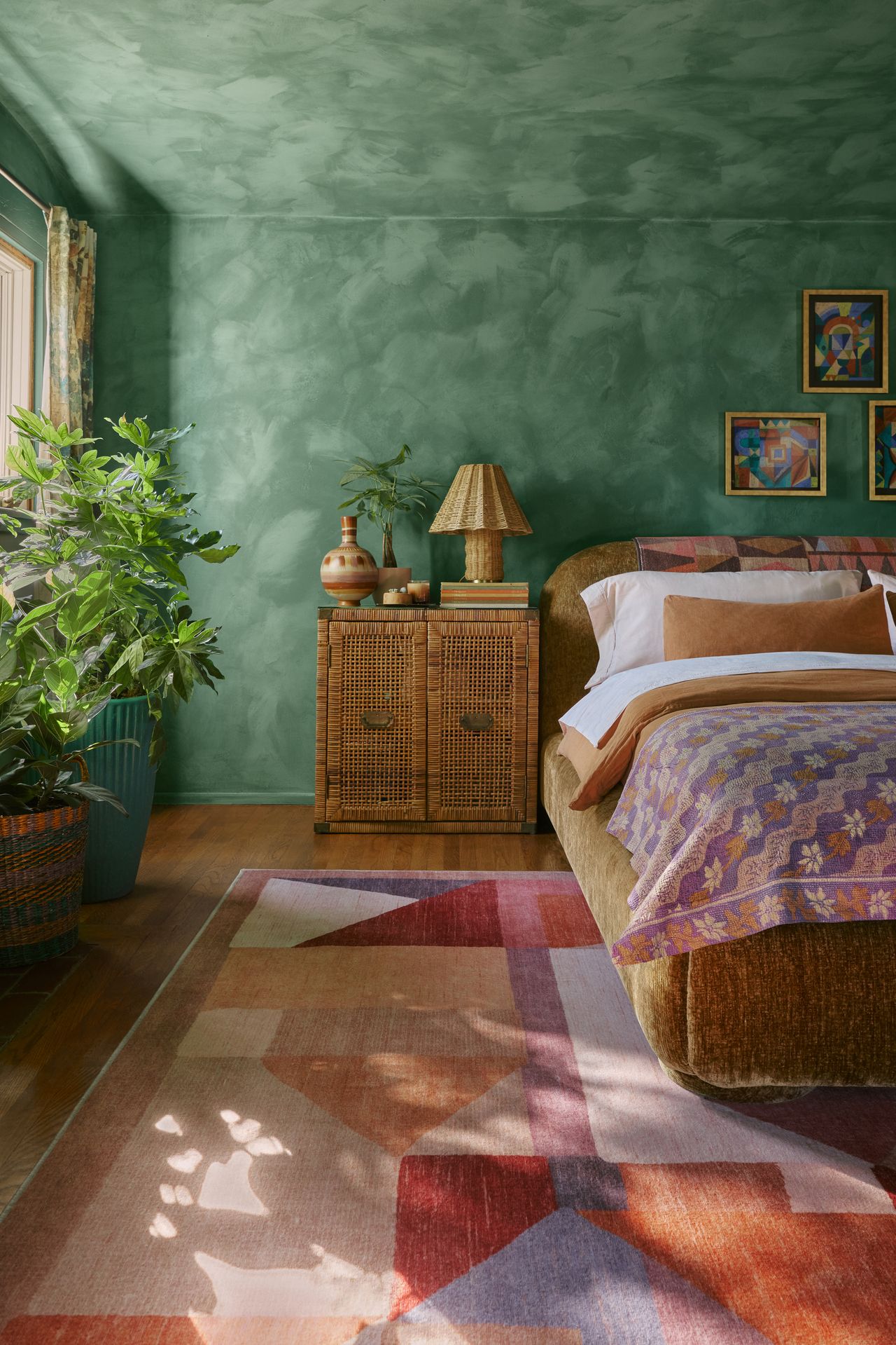 Image of a colorful bedroom with emerald green limewash walls, a warm color geometric rug, an ochre yellow velvet bed, and a rattan bedside table.