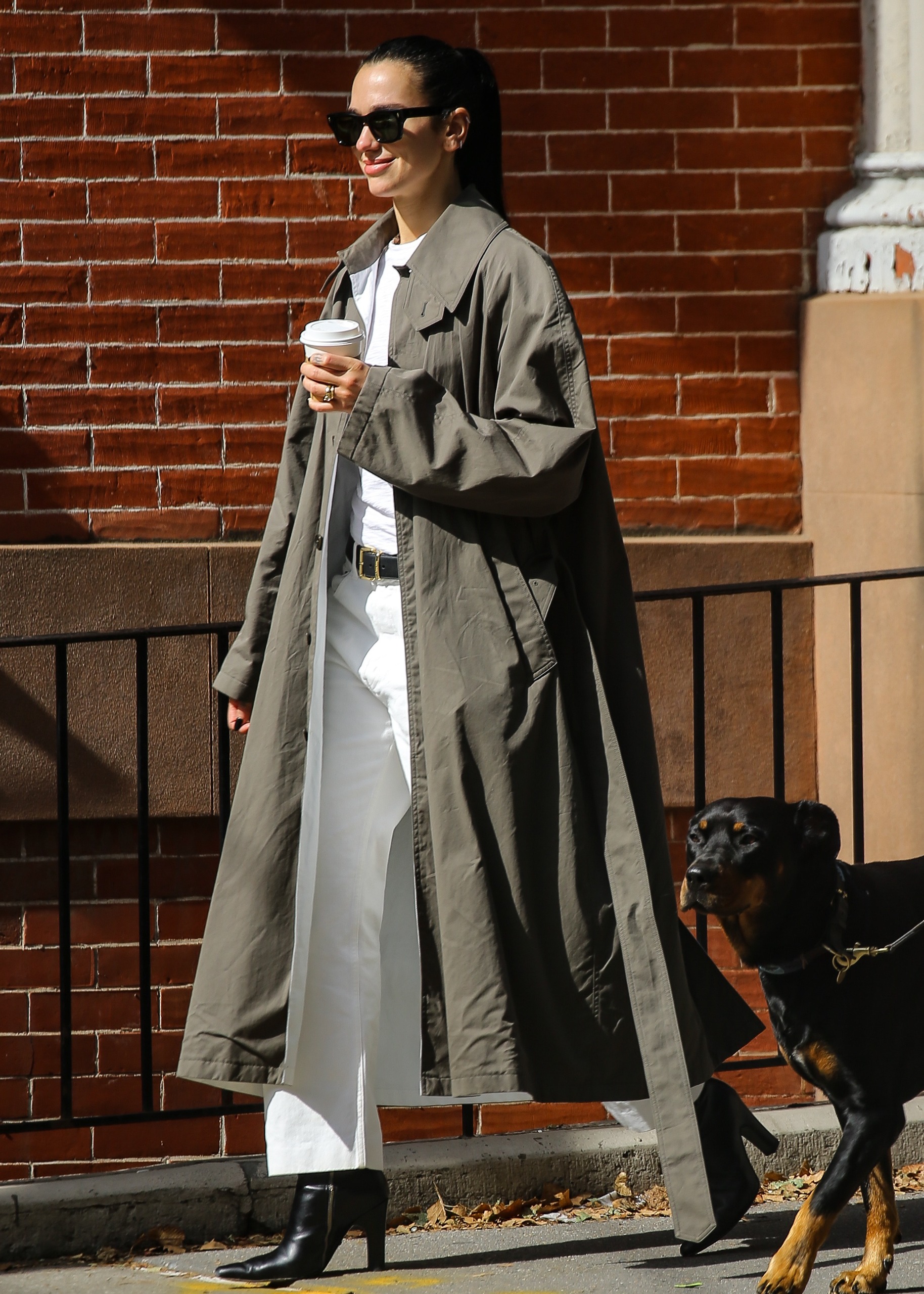 Dua Lipa wears white pants with an anti trend trench coat and black boots while walking with husband Callum Turner