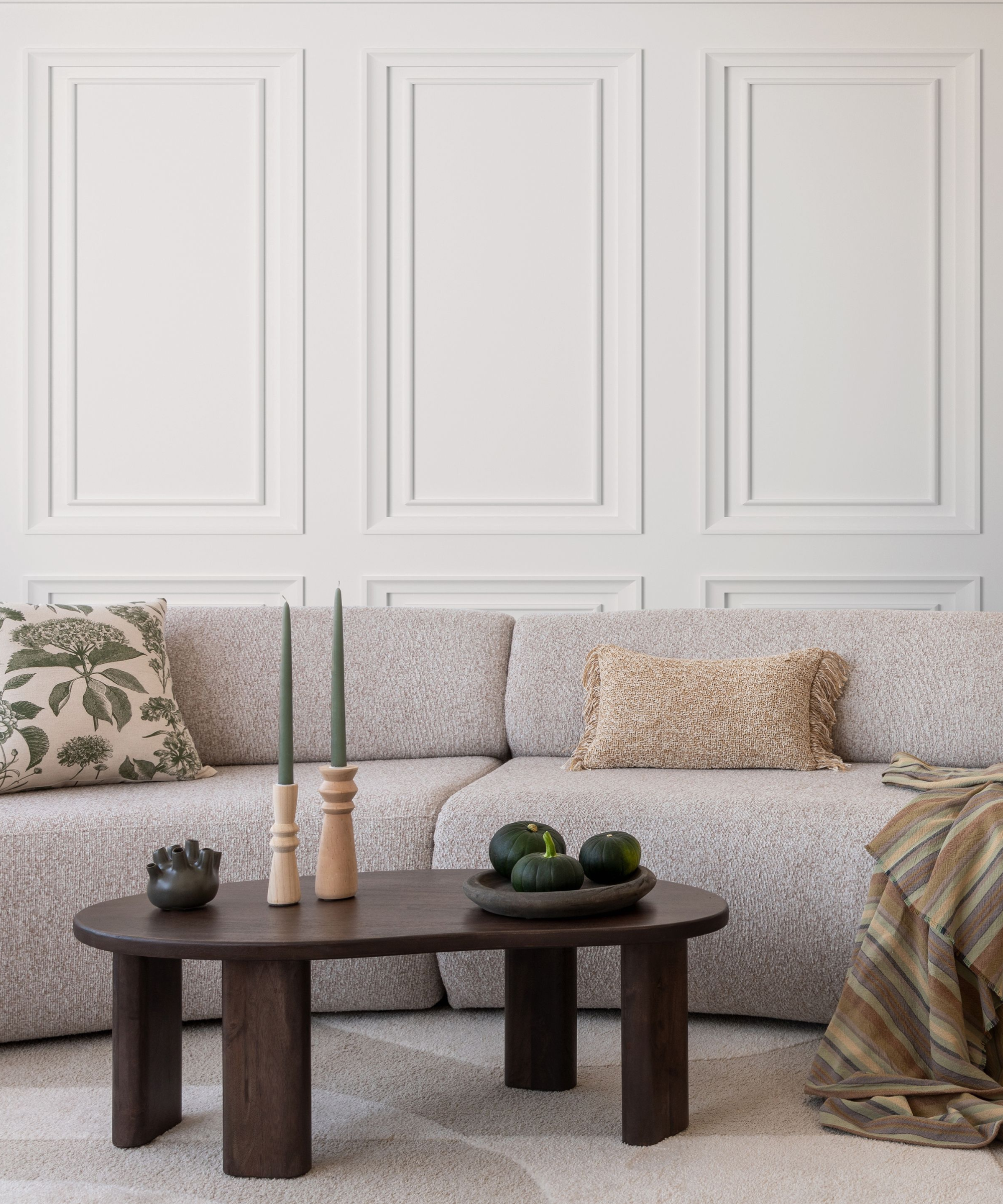 Living room with white raised panelled wall, beige curved sofa, dark brown coffee table and green, beige acessories