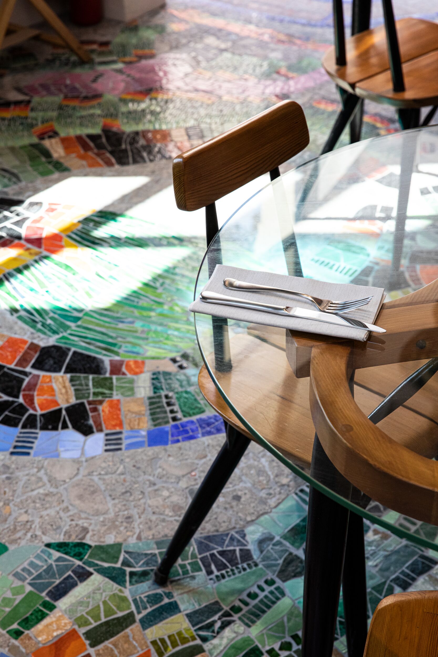 A cafe with glass tables, wooden chairs, and tiled, terrazzo floors in bright hues.