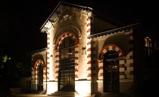 Night time, outside image of the Hotel Ville d'Hiver, Arachon, up lights on the brick building, with redand white arch framed windows, stone steps, shrubs and trees