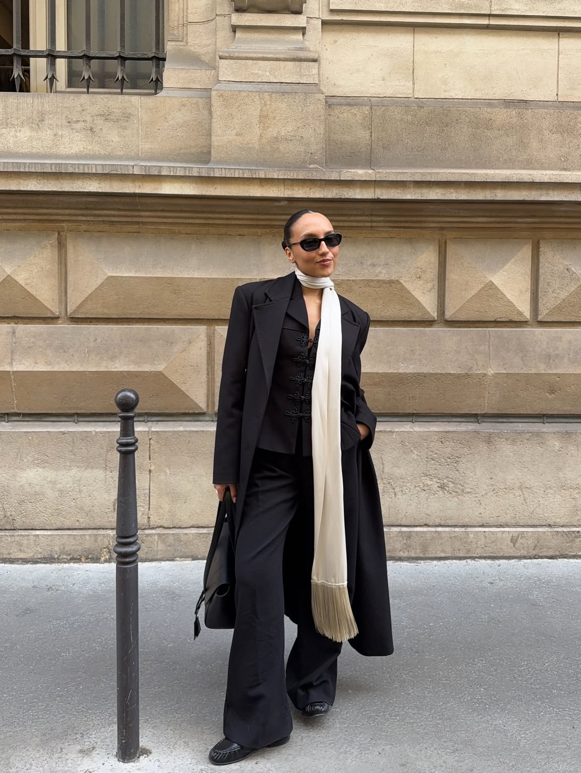 A woman wears a long black coat, black trousers and a white fringe scarf