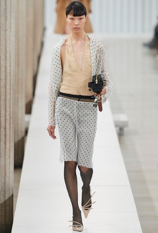 a photo of a model wearing a sheer polka dot top over a nude halter top with a polka dot sheer skirt and tights and nude kitten heels on Miu Miu's fall 2023 runway