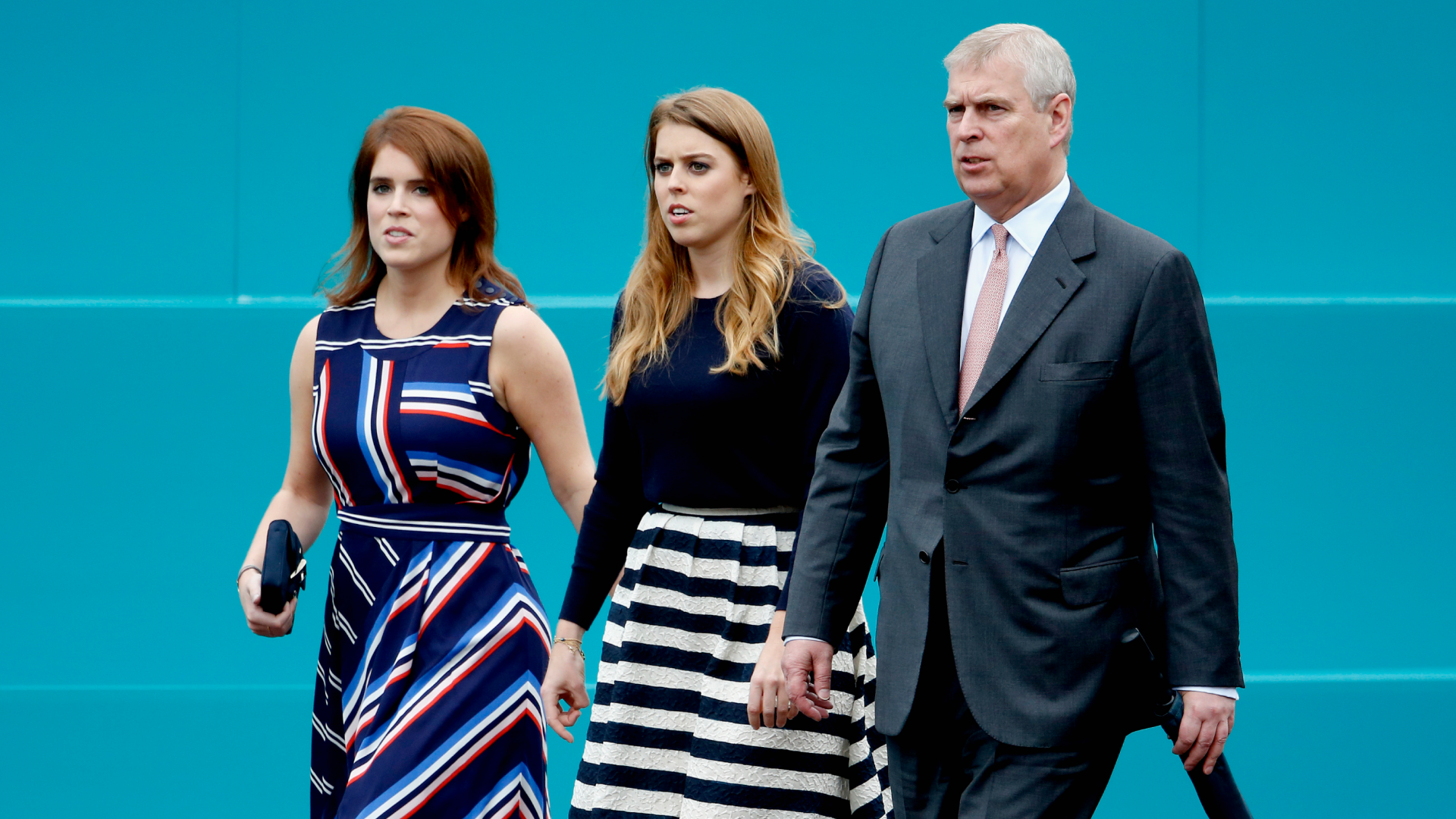 Princess Eugenie, Princess Beatrice and Prince Andrew walking in a row in front of a teal wall