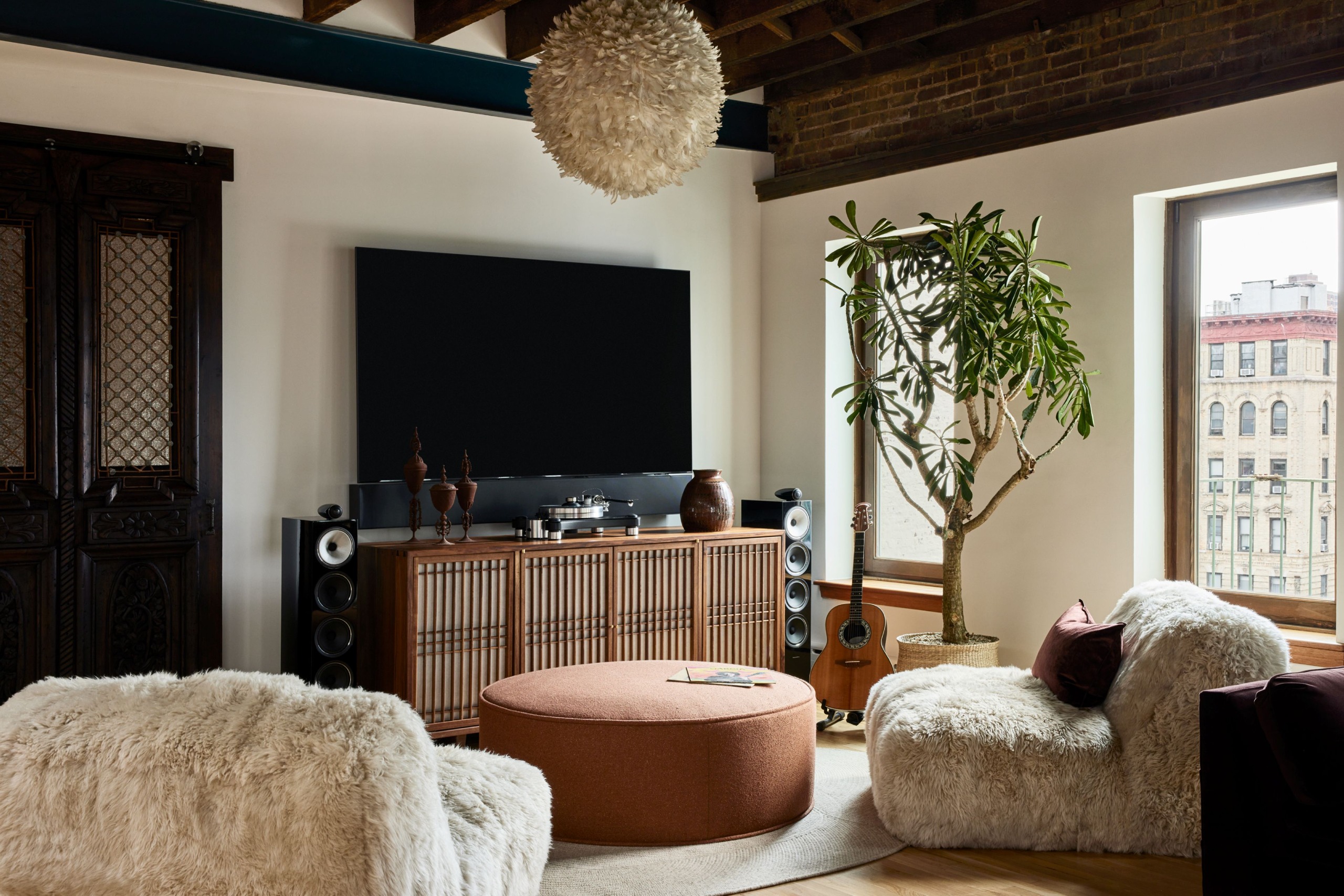 Image of a cozy living room with two fuzzy armless chairs, a round orange ottoman, a wooden console, and a TV with a large speaker system.