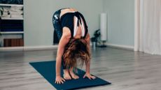 woman performing a rolldown from standing, touching her hands to the floor on an exercise mat and wooden floor.