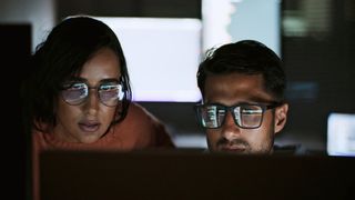 Two cyber workers with medium-dark skin tone, a woman on the left and a man on the right with his face from chin down obscured by a monitor, with code reflected in their glasses implying they work in cybersecurity or software engineering. The room is dark around them, but bright screens can be seen in the background.