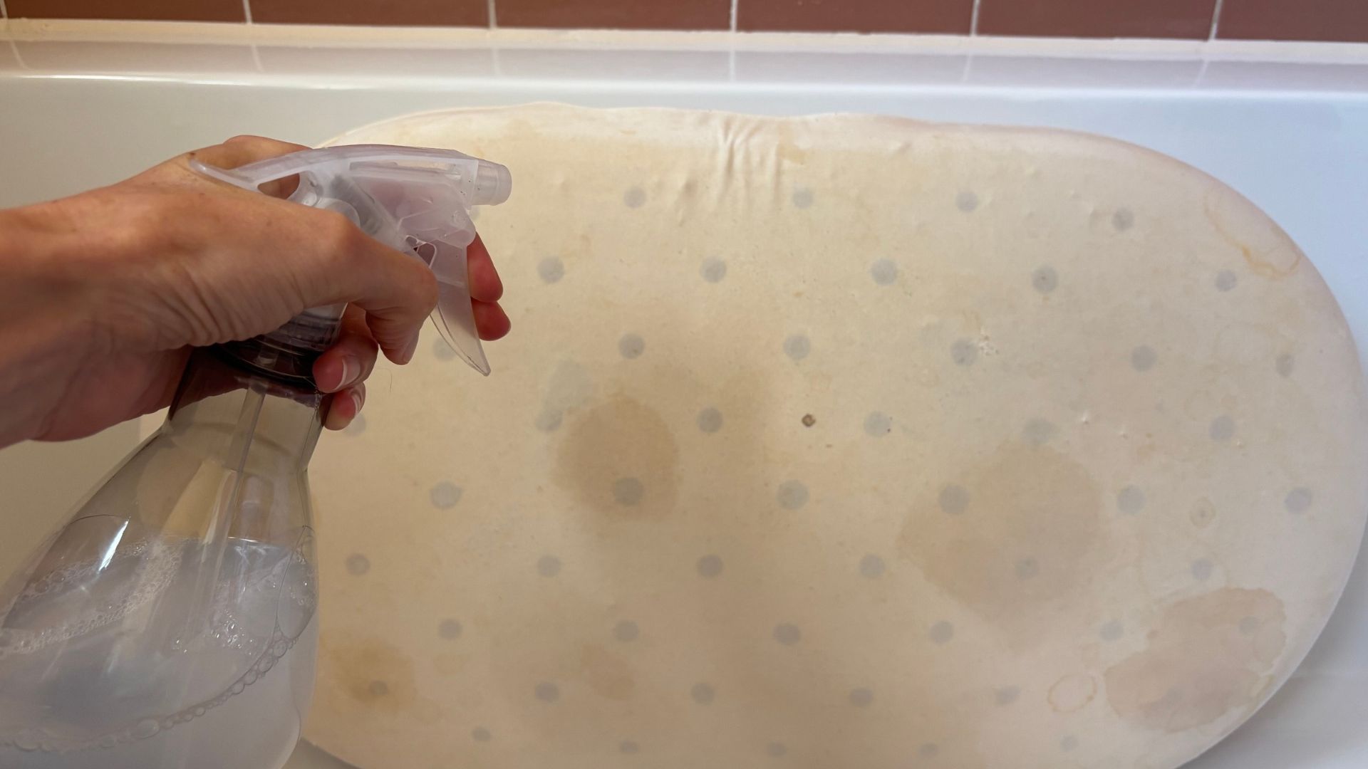 A hand spritzes a stained pillow with a cleaning solution from a spray bottle