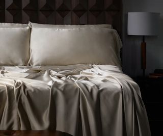 Beige sateen sheets on a bed against a dark bedroom wall.