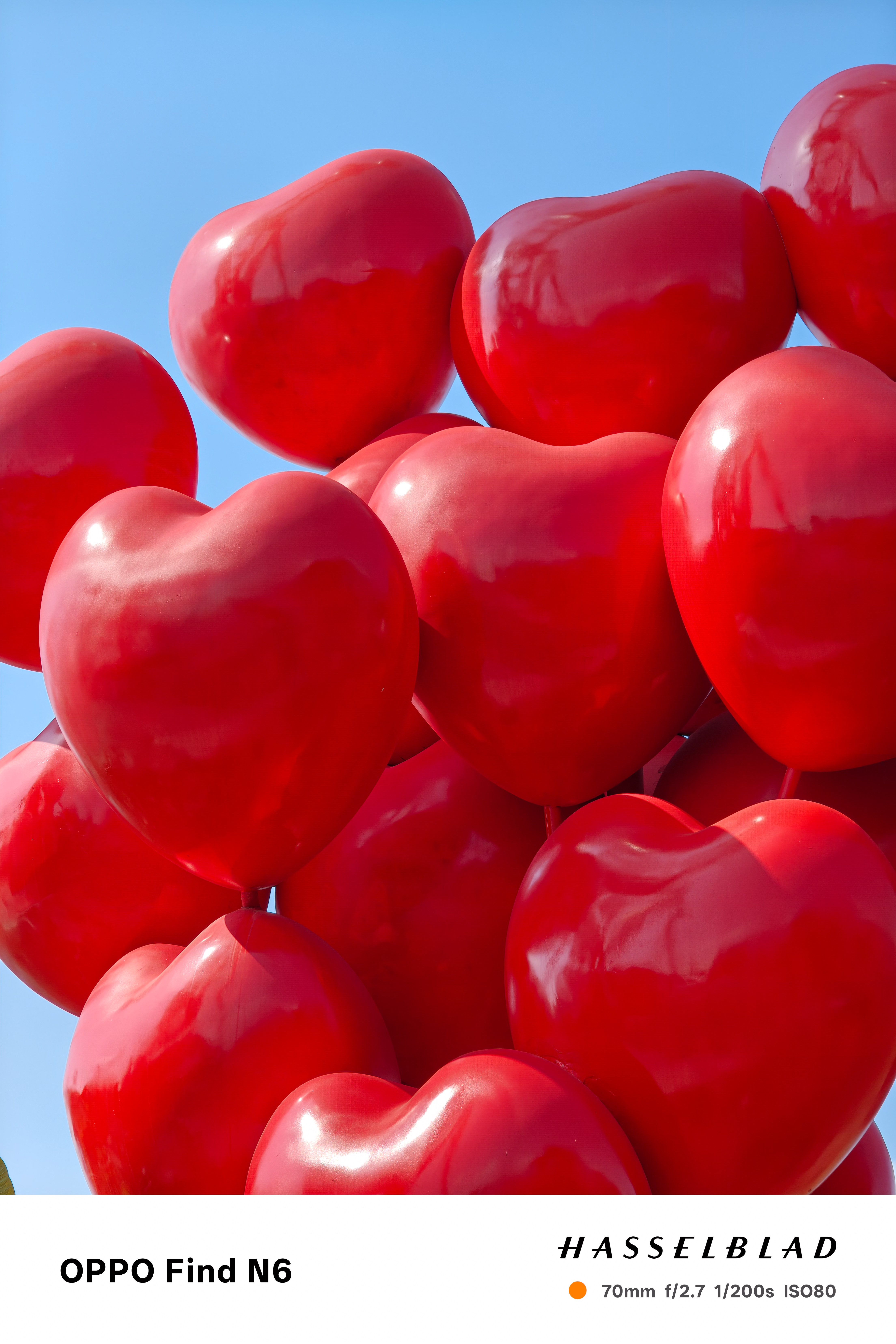 A close-up, high-angle shot of a large cluster of bright red, heart-shaped balloons. The balloons have a glossy, reflective finish that catches the light, packed tightly together against a clear blue sky.