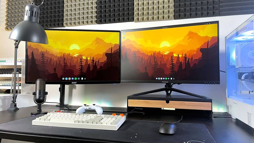 A desk with two monitors, a keyboard, a mouse, a lamp, and a desktop gaming PC on it showing a Linux home screen